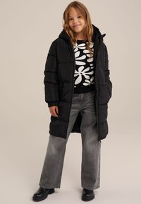 Černý vycpaný dlouhý kabát s kapucí, nošený přes vzorovaný černobílý svetr. V páru s světlými šedými širokými kalhotami a černými kotníkovými botami.