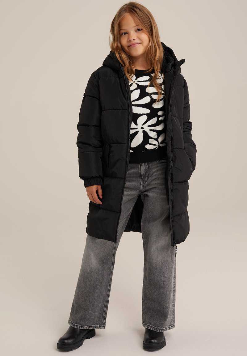 Černý vycpaný dlouhý kabát s kapucí, nošený přes vzorovaný černobílý svetr. V páru s světlými šedými širokými kalhotami a černými kotníkovými botami.