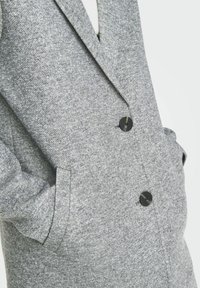 Manteau double boutonnage en gris texturé avec des boutons noirs et des poches latérales, présentant un design de col taillé et un tissu tissé doux.