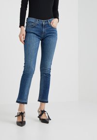 Vaqueros de mezclilla azul con un diseño ajustado, cintura de altura media y dobladillos deshilachados en los tobillos, combinados con tacones negros de punta fina con acentos de cinta.