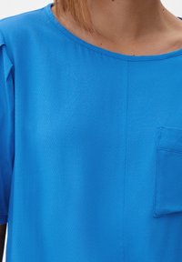 Nahaufnahme einer Person, die ein leuchtend blaues, strukturiertes Hemd mit einer kleinen Brusttasche und kurzen Ärmeln trägt.