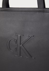 Černá kožená taška přes rameno s vyraženým logem "CK", hladkým povrchem a pevným uchy. Design je minimalistický a elegantní.