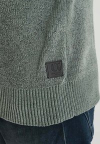 Nahaufnahme des Saums eines grünen Strickpullovers mit geripptem Rand und einem schwarzen rechteckigen Markenpatch, getragen über einer dunkelblauen Jeans.
