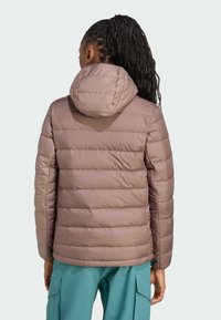 Donna con lunghe trecce che indossa un giubbotto imbottito mauve con cappuccio e pantaloni cargo color teal, vista da dietro su uno sfondo chiaro e semplice.