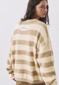 Kremasto-rjav trakast pulover z mehkimi vlakni in sproščenim krojem, ki ima kvadratno etiketo na hrbtni strani z napisoma "Od 2018" in "Ustvarjeno za življenje".