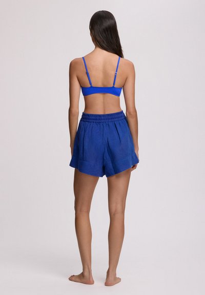 Γυναίκα όρθια ξυπόλυτη, με το πρόσωπο στραμμένο προς τα πίσω, φοράει μπλε κορυφή κομμένη με λεπτές τιράντες και χαλαρά μπλε σορτς, μπροστά σε απλό φόντο.