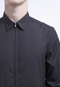 Chemise noire à fermeture éclair avec col pointu, manches longues, texture lisse et design minimaliste. La fermeture éclair descend verticalement sur le devant.