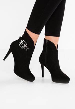 Schwarze Wildleder-Stiefeletten mit hohem Stiletto-Absatz, asymmetrischem Schnitt, seitlichem Reißverschluss und einer dekorativen Schnalle mit Ösen.