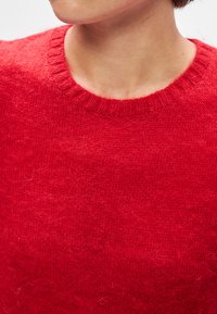 Piros kötött pulóver kerek nyakkivágással. Az anyag puha, textúrált felületű, enyhén bolyhos megjelenéssel.
