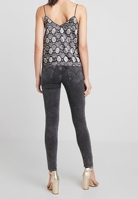 Femme portant un débardeur en imprimé serpent, un jean skinny noir et des sandales à talons blocs dorés, se tenant de dos.