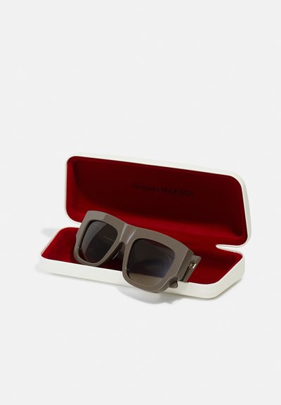 Ochelari de soare gri supradimensionați într-un capac alb cu interior roșu. Prezintă rame pătrate îndrăznețe și lentile întunecate. Brandingul este vizibil în interiorul capacului.