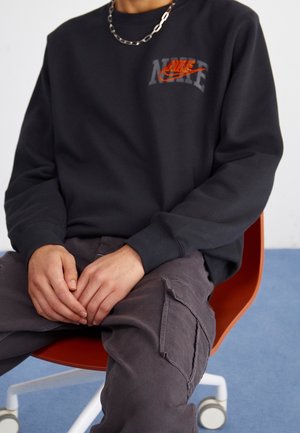 Person sidder på orange stol iført sort Nike-sweatshirt med orange logo og grå cargobukser, hænderne hvilende på skødet.