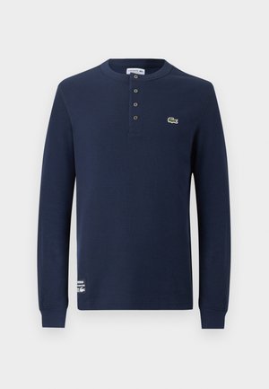 Marineblå langærmet henley-shirt med ribbet tekstur, fire knapper, Lacoste krokodillelogo på brystet og mærkelabel nær kanten.