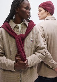 Bézs kockás gombos ing mellzsebbel, hozzá párosítva egy bordó, kötött mintás pulóverrel, amelyet a nyak köré kötöttek; a modell bordó sapkát visel.