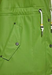 Vihreä vedenpitävä takki, jossa sileä pinta, vino tasku, valkoiset kiristysnauhat ja pyöreät, merkkikoristeiset napit.