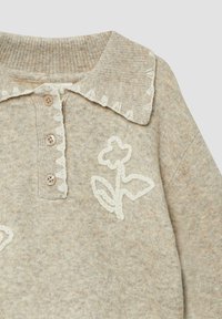 Beige gebreide trui met een zachte textuur, voorzien van een gegolfde kraag, drie knopen en een geborduurd bloemenmotief in het wit.