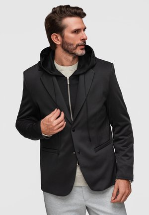 Homme avec une barbe taillée portant un blazer noir par-dessus un sweat à capuche zippé noir et un pantalon gris clair, regardant vers sa gauche.