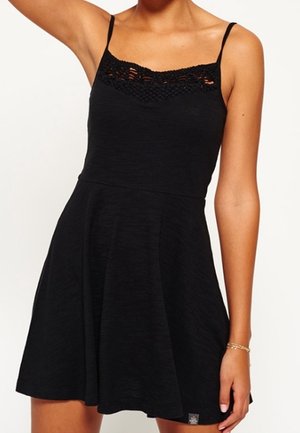 Femme portant une robe noire sans manches avec un détail en dentelle le long du décolleté et des fines bretelles, debout avec un bras levé.