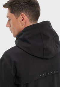 Veste à capuche noire avec une texture lisse, présentant un design à dos superposé et des coutures décoratives blanches le long de la couture d'épaule.