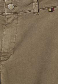 Tessuto denim beige con una fine texture a spina di pesce, caratterizzato da un bottone metallico e un piccolo etichetta colorata a strisce sulla cintura.