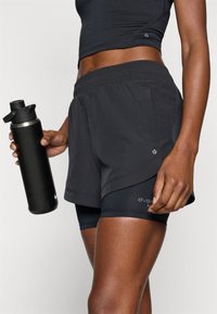 Pantalones cortos atléticos negros con un diseño en capas sobre pantalones ciclistas ajustados. Presenta un logo plateado y bolsillos. La modelo sostiene una botella de agua negra.