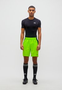 Αθλητής που στέκεται και κοιτάζει μπροστά, φοράει μαύρο συμπιεστικό μπλουζάκι, αθλητικά σορτς σε νέον πράσινο χρώμα, μαύρες κάλτσες Adidas μέχρι το γόνατο και μαύρα παπούτσια με αγκράφες.