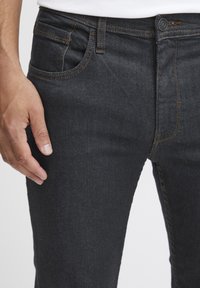 Mörka denimjeans med orange sömmar, med en framficka och en figurnära passform. Närbild på en hand som vilar på låret.
