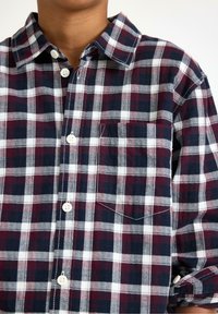 Camicia in flanella a quadretti con quadrati blu navy, borgogna e bianchi, con bottoni bianchi davanti e una piccola tasca sul petto. Tessuto morbido.