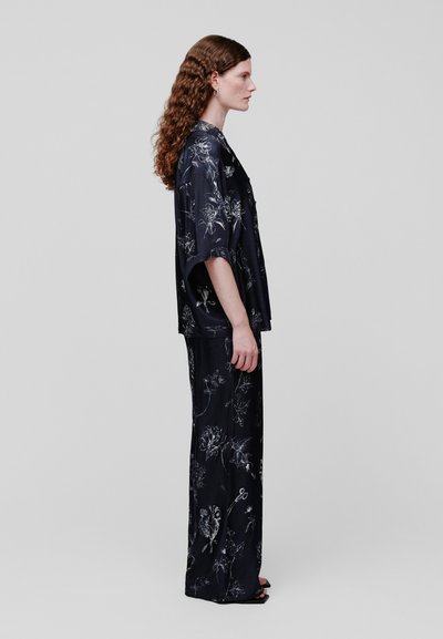 Femme aux longs cheveux bouclés, debout de profil, portant une chemise ample à manches courtes à motif floral foncé et un pantalon large assorti avec des sandales noires.