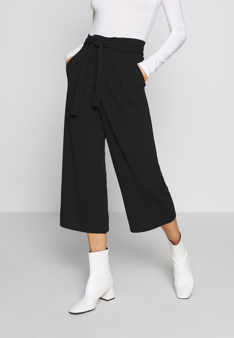 Pantalons noirs à jambes larges avec taille haute, détail de ceinture et poches latérales, associés à des bottines blanches à bout carré et talon bloc.
