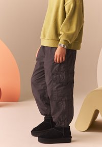 Pantaloni cargo in tessuto grigio scuro con polsini elasticizzati, abbinati a stivali alla caviglia neri in suede. Il maglione è verde oliva con polsini a righe.