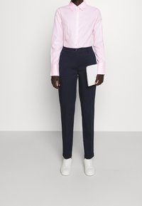 Chemise rose clair à manches longues avec col et boutons, associée à un pantalon slim bleu marine et des baskets blanches, tenant une tablette blanche.