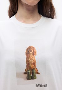 Γυναίκα που φοράει άσπρο πουκάμισο με μια καφέ σκυλίτσα να κάθεται πίσω από δύο πράσινα μπότες Barbour, με το κείμενο "BARBOUR" από κάτω.