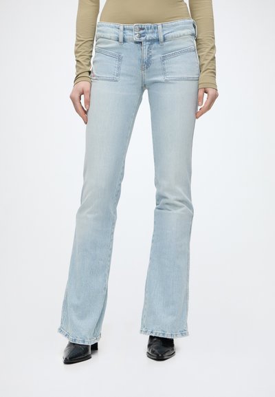 Personne portant un jean évasé bleu clair avec des poches plaquées à l'avant et des bottes noires à bouts pointus sur un fond blanc uni.