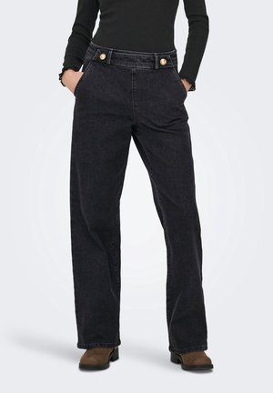 Personne portant un jean noir taille haute à jambes larges avec des boutons dorés, mains dans les poches, haut noir à manches longues et bottes marron.