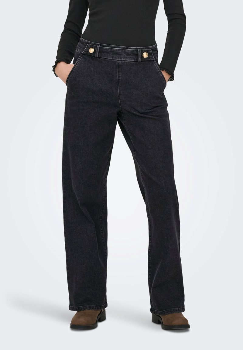 Personne portant un jean noir taille haute à jambes larges avec des boutons dorés, mains dans les poches, haut noir à manches longues et bottes marron.