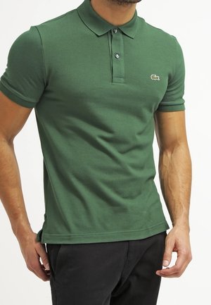 Mann trägt ein eng anliegendes grünes Poloshirt mit kleinem Krokodil-Logo auf der Brust und schwarze Hose vor weißem Hintergrund.