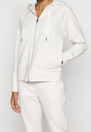 Kobieta ubrana w biały hoodie z suwakiem i markowymi sznurkami oraz pasujące białe spodnie dresowe, pozująca z jedną ręką w kieszeni.