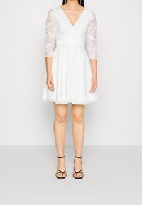 Weißes Kleid mit V-Ausschnitt und dreiviertellangen Spitzenärmeln, ausgestellter Rock, strukturierter Spitzenoberteil, kombiniert mit schwarzen Riemchen-Heels.