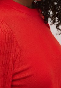 Close-up van een persoon die een levendige rode gebreide trui draagt met een gedetailleerde ribstructuur op de mouwen en zichtbaar krullend donker haar.