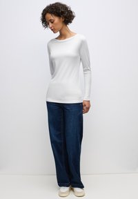 Langärmliges, Rundhals-Weißes Oberteil mit glatter Textur, kombiniert mit weit geschnittenen dunkelblauen Jeans und weißen Sneakern. Einfaches und lässiges Outfit.