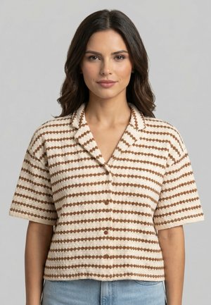 Donna con capelli scuri che indossa una camicia beige e marrone a righe con maniche corte e bottoni, e jeans azzurri, in piedi davanti a uno sfondo grigio.