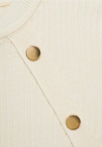 Gros plan sur un vêtement en tissu côtelé crème avec une bride diagonale présentant deux boutons ronds en métal doré.