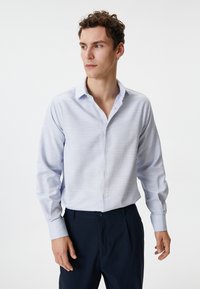Lichtblauw overhemd met een subtiele gestreepte textuur, lange mouwen en een kraag. Draagt bij donkerblauwe broek.