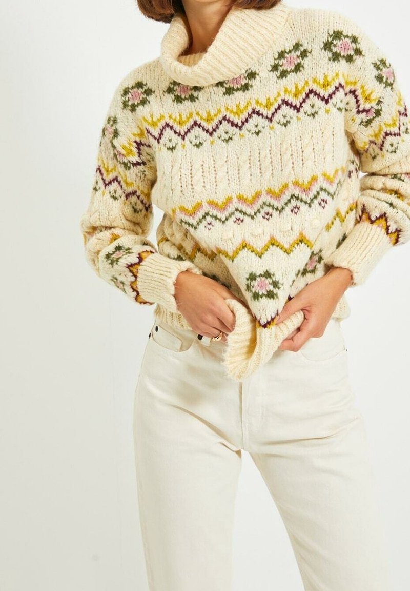 Pull en tricot crème avec motifs floraux et en zigzag porté avec un pantalon blanc, mains ajustant l'ourlet du pull à la taille.