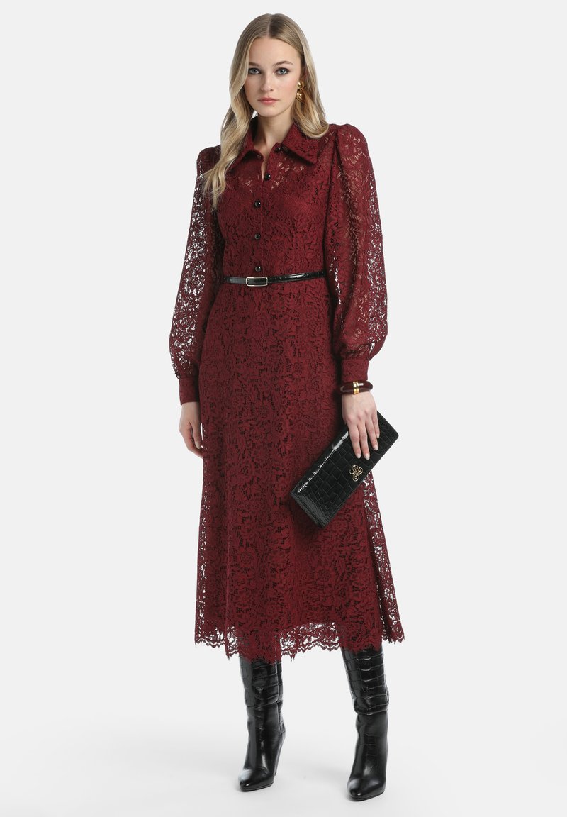 Burgunderfarbenes Spitzenkleid mit langen Puffärmeln, Knopfverschluss vorne und taillierter Taille, kombiniert mit einer schwarzen Clutch und knielangen schwarzen Stiefeln.
