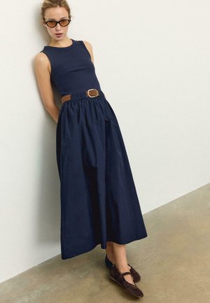 Ženska, oblečena v brezrokavno temno modro obleko z rjavim pasom, temno rjave balerinke in sončna očala, ki se naslanja na belo steno v notranjosti.