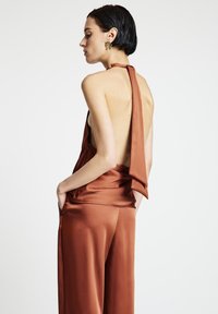 Satin-Halftertop in Rostfarbe mit offenem Rücken, ausgestattet mit einem fließenden Fall und breiten Trägern. Hose aus passendem Stoff mit Taschen.