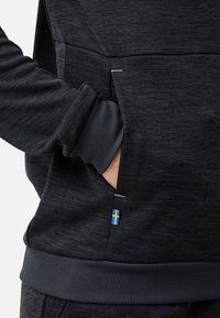 Hand einer Person in der Tasche eines dunkelgrauen strukturierten Sweatshirts mit einem schwedischen Flaggen-Etikett nahe der Taschennähte.