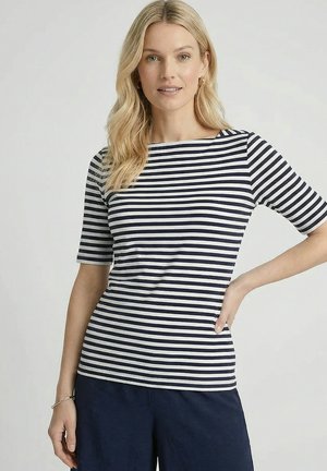 Vrouw met blond haar die een marineblauw-wit gestreepte top met korte mouwen en marineblauwe broek draagt, poseert met één hand op haar heup.